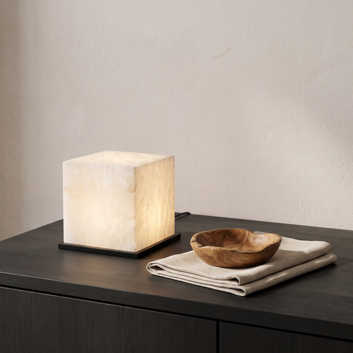 Alabaster table lamp glowing on a dark wood surface beside a carved wood bowl